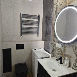 Lucas - Nowoczesna łazienka po remoncie: czarna armatura, umywalka z szafką, toaleta, grzejnik drabinkowy i pralka na tle płytek w dwóch wzorach.