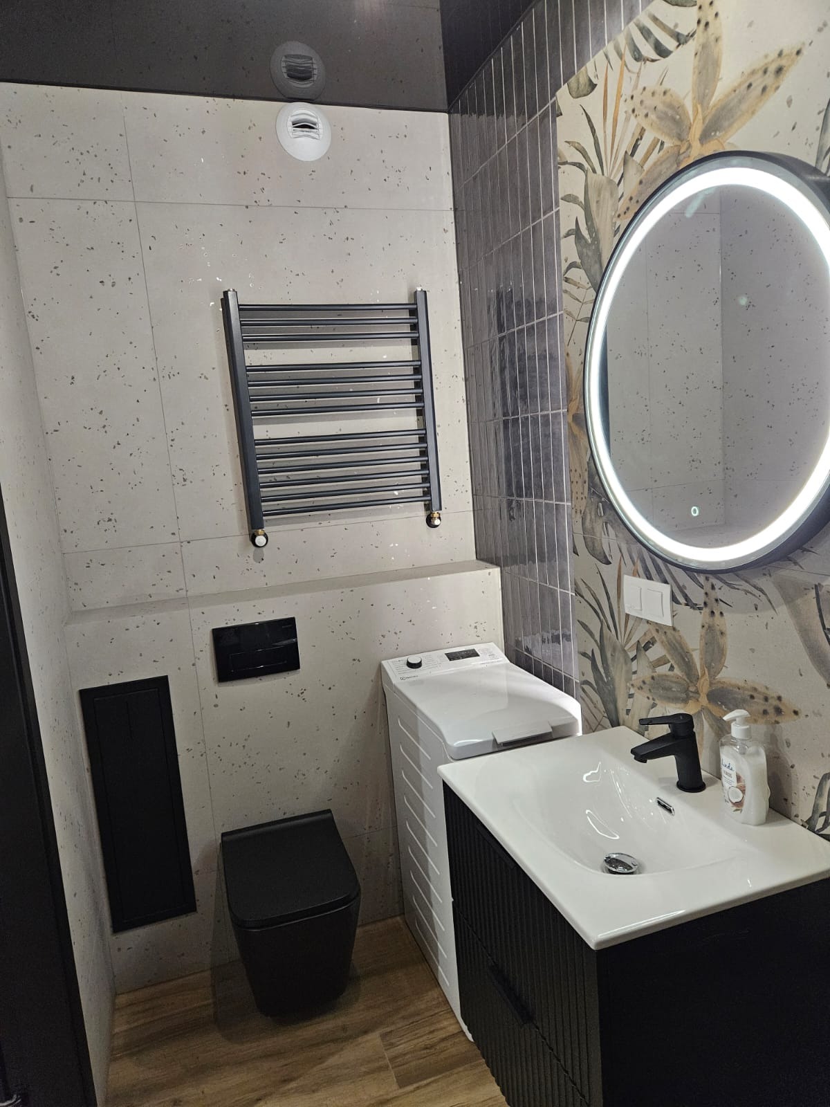 Nowoczesna łazienka z czarną armaturą, umywalką na szafce, podświetlanym lustrem i grzejnikiem drabinkowym na tle płytek w odcieniach szarości i beżu.