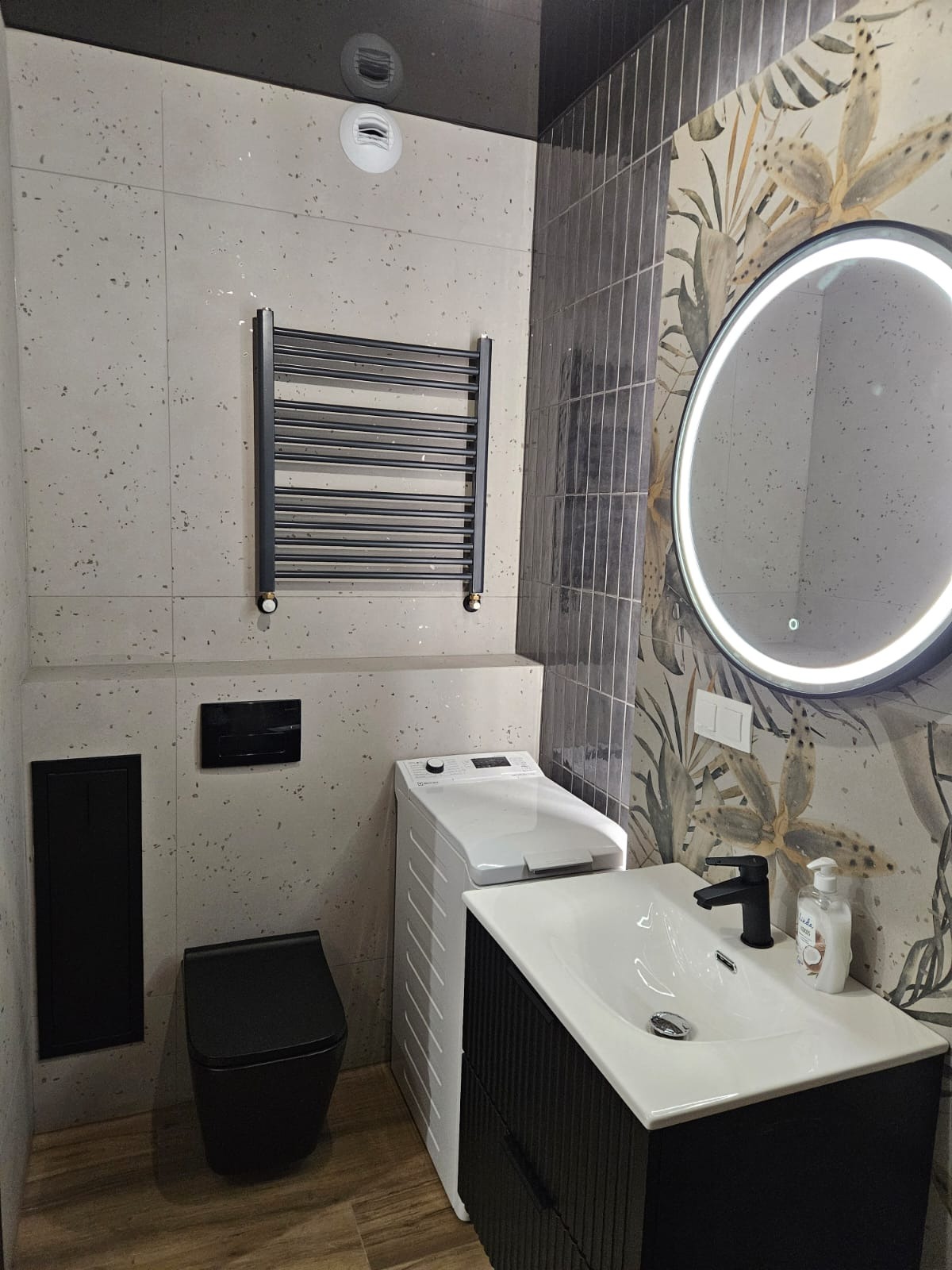 Nowoczesna łazienka po remoncie: czarna armatura, umywalka z szafką, toaleta, grzejnik drabinkowy i pralka na tle płytek w dwóch wzorach.