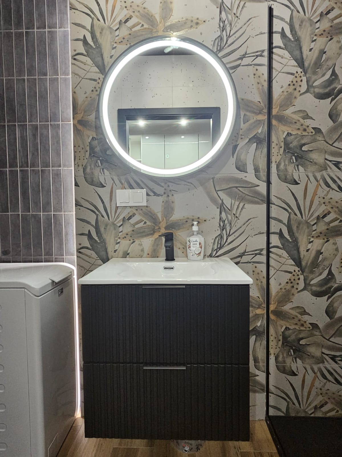 Nowoczesna łazienka: umywalka z szafką w kolorze ciemnego drewna, okrągłe lustro z podświetleniem LED i płytki w tropikalne wzory. Minimalistyczny design.