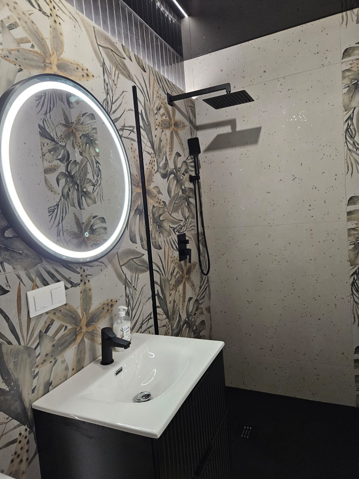 Nowoczesna łazienka z umywalką, okrągłym lustrem z podświetleniem LED i deszczownicą. Ściany wyłożone płytkami z motywem roślinnym i jasnymi płytkami.