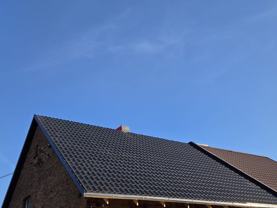 Ciemny, nowy dach z blachodachówki na ceglanym domu pod błękitnym niebem. Widoczne rynny i komin. Ujęcie z dołu, podkreślające geometrię dachu.