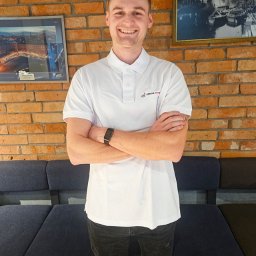 SEWERYN SZKLARZ - Uśmiechnięty mężczyzna w białej koszulce polo z logo firmy sprzątającej, stoi na tle ceglanej ściany i ciemnej sofy. Na ścianie wiszą dwa obrazy w ramkach.