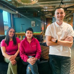 SEWERYN SZKLARZ - Zespół sprzątający w biurze: dwie kobiety siedzą na kanapie, mężczyzna stoi obok z założonymi rękami. W tle stół bilardowy i duże okno.