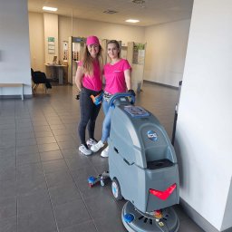 SEWERYN SZKLARZ - Dwie uśmiechnięte kobiety w różowych koszulkach stoją obok szorowarki do podłóg w przestronnym, jasnym wnętrzu z szarą podłogą z płytek.