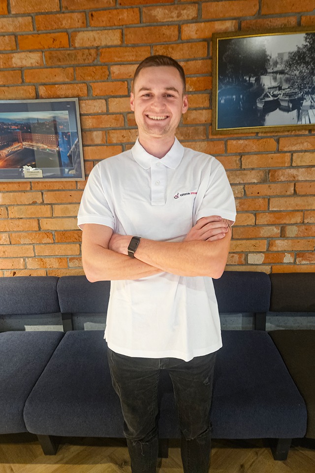 Uśmiechnięty mężczyzna w białej koszulce polo z logo firmy sprzątającej, stoi na tle ceglanej ściany i ciemnej sofy. Na ścianie wiszą dwa obrazy w ramkach.