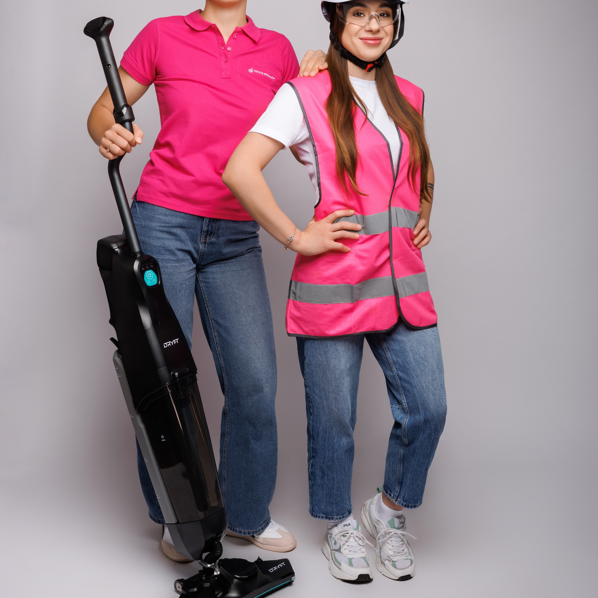 Dwie kobiety w różowych koszulkach i jeansach, jedna w kasku i kamizelce odblaskowej, prezentują czarny mop parowy Dryft na szarym tle. Uśmiechnięta pracownica.