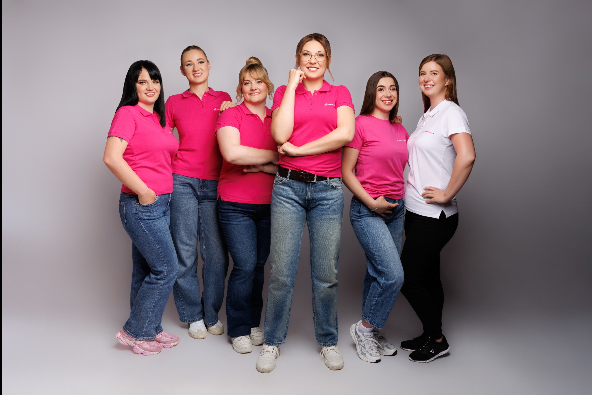 Zespół sprzątający: sześć uśmiechniętych kobiet w różowych i białych koszulkach polo, stojących na szarym tle. Profesjonalny wygląd, schludne ubrania.