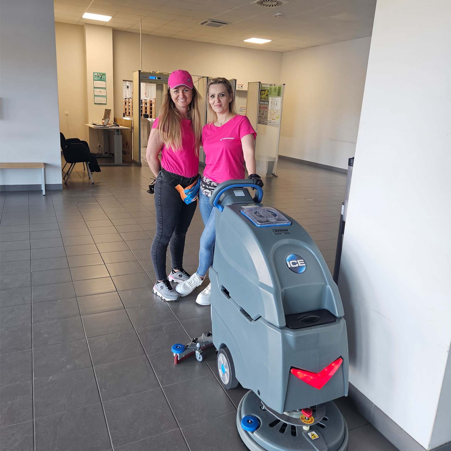 Dwie uśmiechnięte kobiety w różowych koszulkach stoją obok szorowarki do podłóg w przestronnym, jasnym wnętrzu z szarą podłogą z płytek.