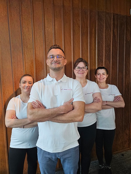 Zespół sprzątający w białych koszulkach polo z logo firmy, stojący na tle drewnianej ściany. Uśmiechnięci, z założonymi rękami, gotowi do pracy.