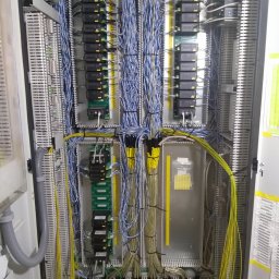Sławomir Troszczynski - Rozdzielnica elektryczna z licznymi przewodami w kolorach niebieskim i żółtym, modułami sterującymi i listwami zaciskowymi. Widoczne elementy instalacji wewnątrz szafy.