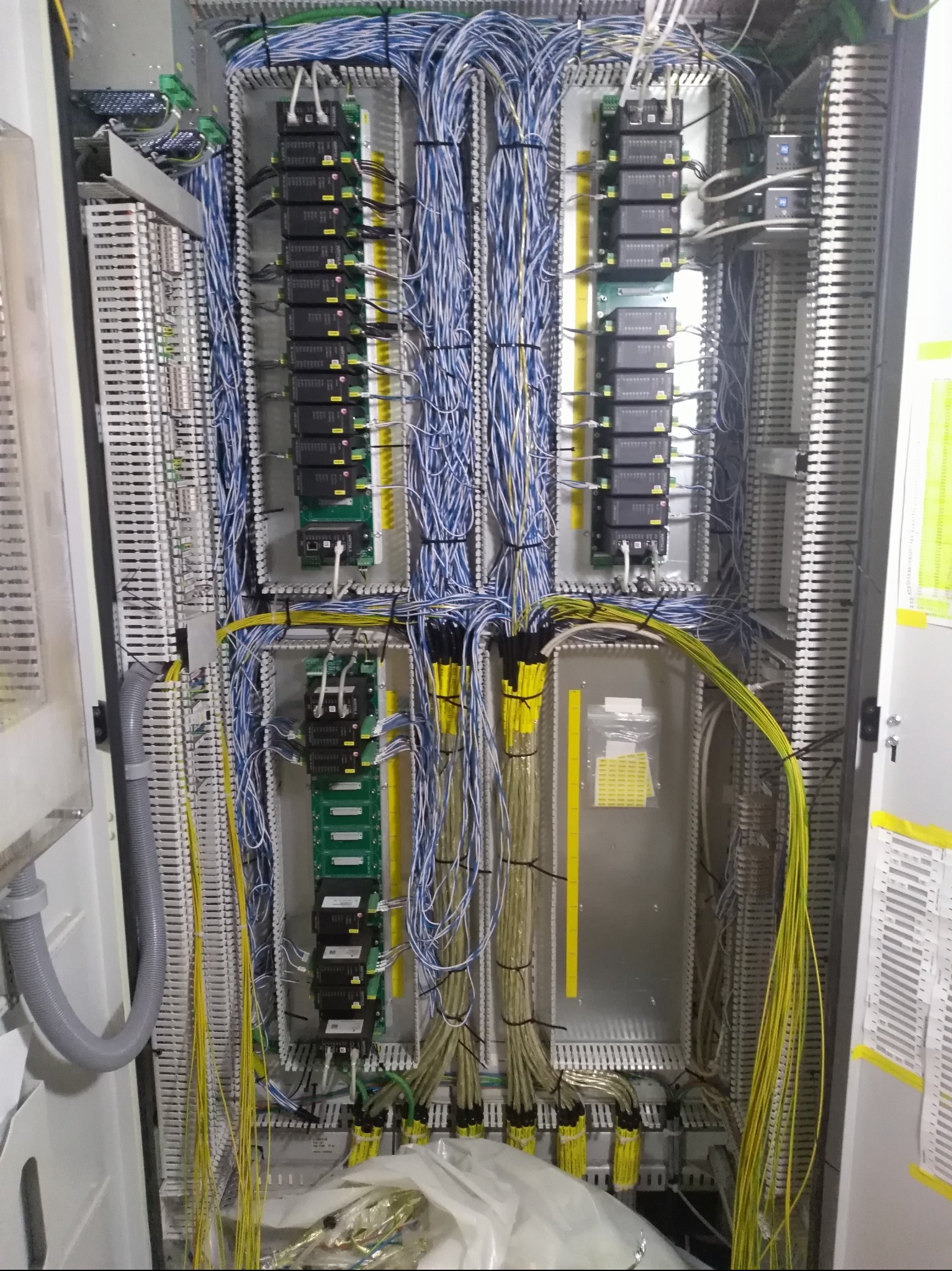 Rozdzielnica elektryczna z licznymi przewodami w kolorach niebieskim i żółtym, modułami sterującymi i listwami zaciskowymi. Widoczne elementy instalacji wewnątrz szafy.