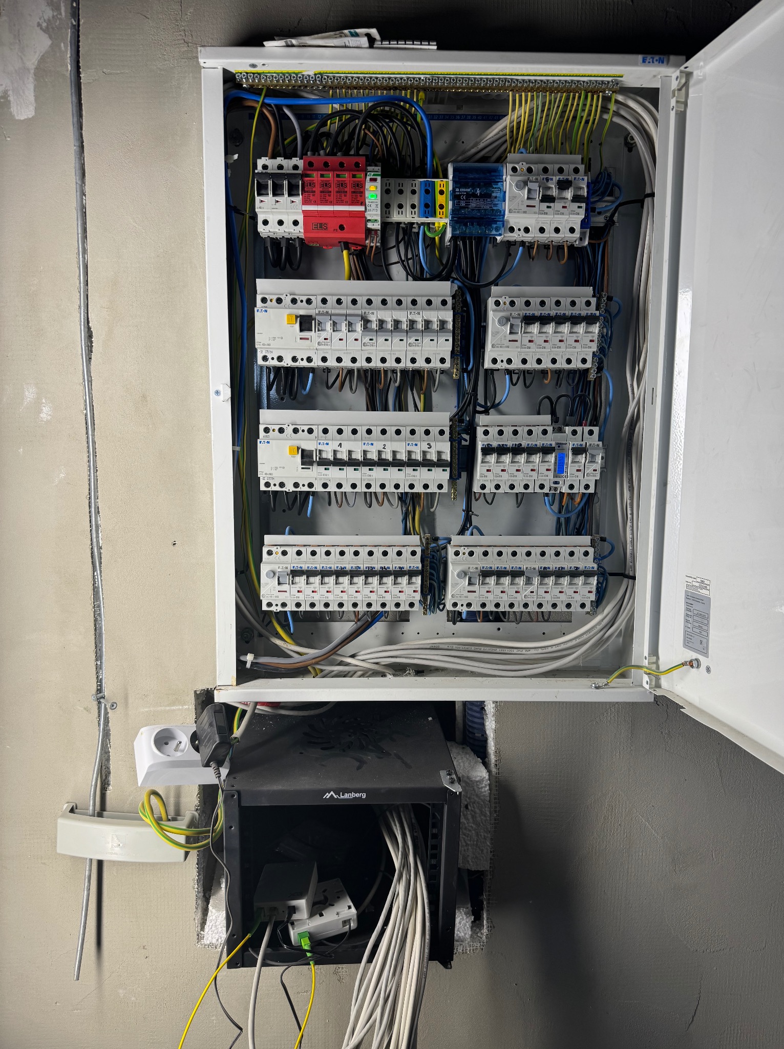 Rozdzielnica elektryczna z zabezpieczeniami różnicowoprądowymi i nadprądowymi, widoczne kable w kolorach: niebieskim, żółto-zielonym, czarnym. Obok puszka instalacyjna.