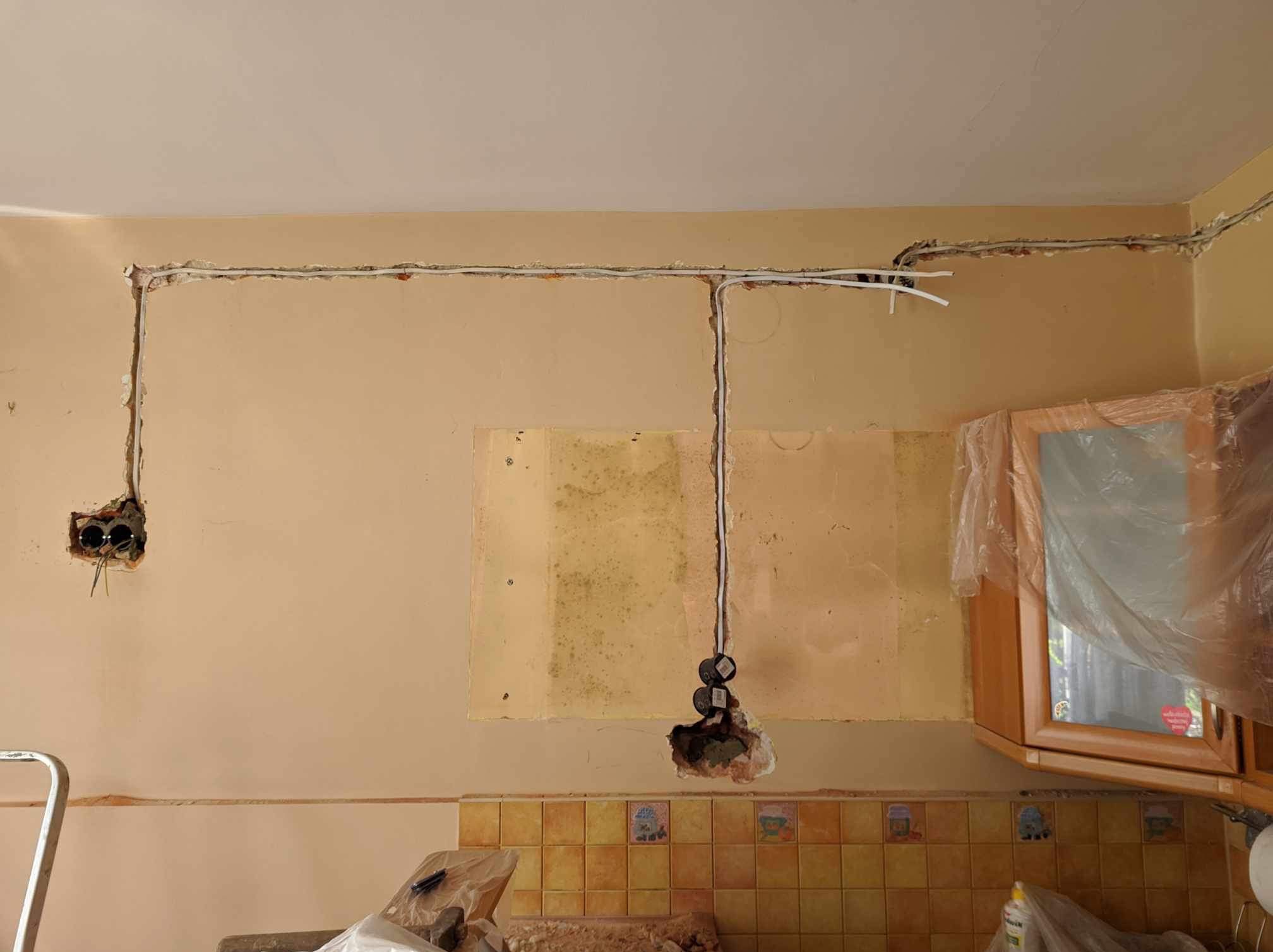 Wnętrze w trakcie remontu: widoczne kable elektryczne prowadzone po ścianie, puszki instalacyjne i fragmenty instalacji podtynkowej. Ściany w odcieniach beżu i żółci.