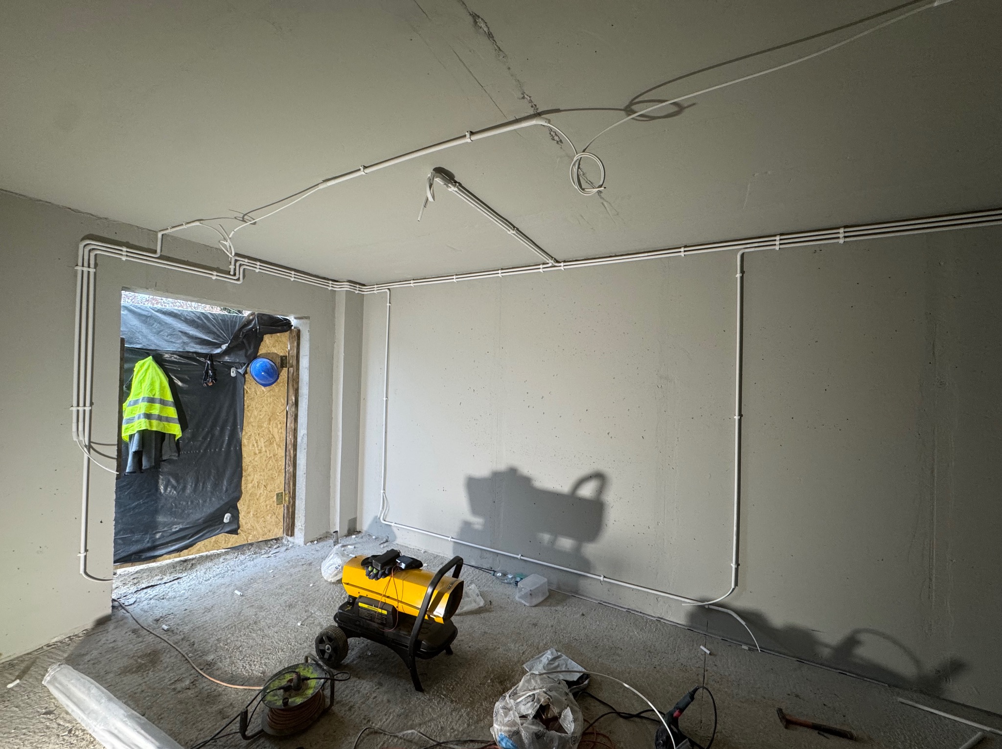 Białe rury instalacji elektrycznej ułożone na ścianach i suficie surowego wnętrza. Widoczny nagrzewnica, narzędzia i odzież robocza. Etap prac instalacyjnych.