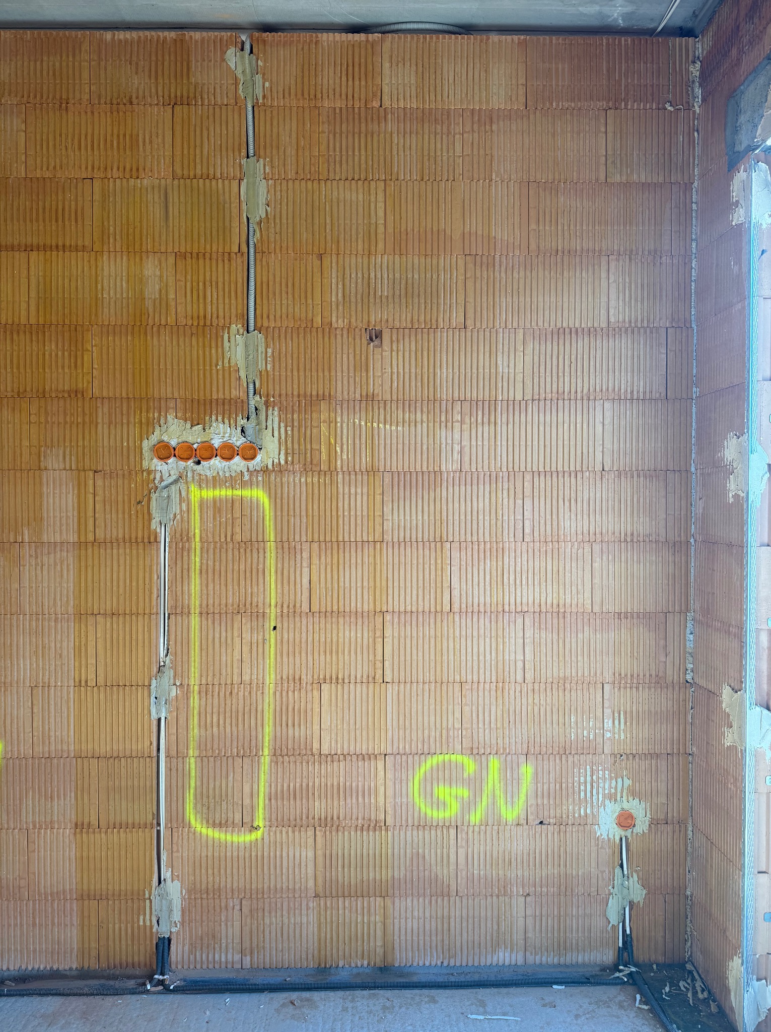 Ściana z pustaków z bruzdami wypełnionymi instalacją elektryczną. Widoczne puszki pod gniazdka i przewody w peszlach. Na ścianie żółte oznaczenia.