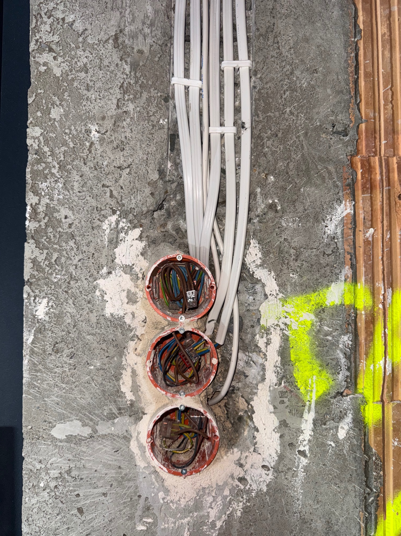 Surowa ściana z trzema okrągłymi puszkami instalacyjnymi, z których wychodzą białe przewody. Widoczne kable wewnątrz puszek i fragment ściany z cegły z żółtą farbą.