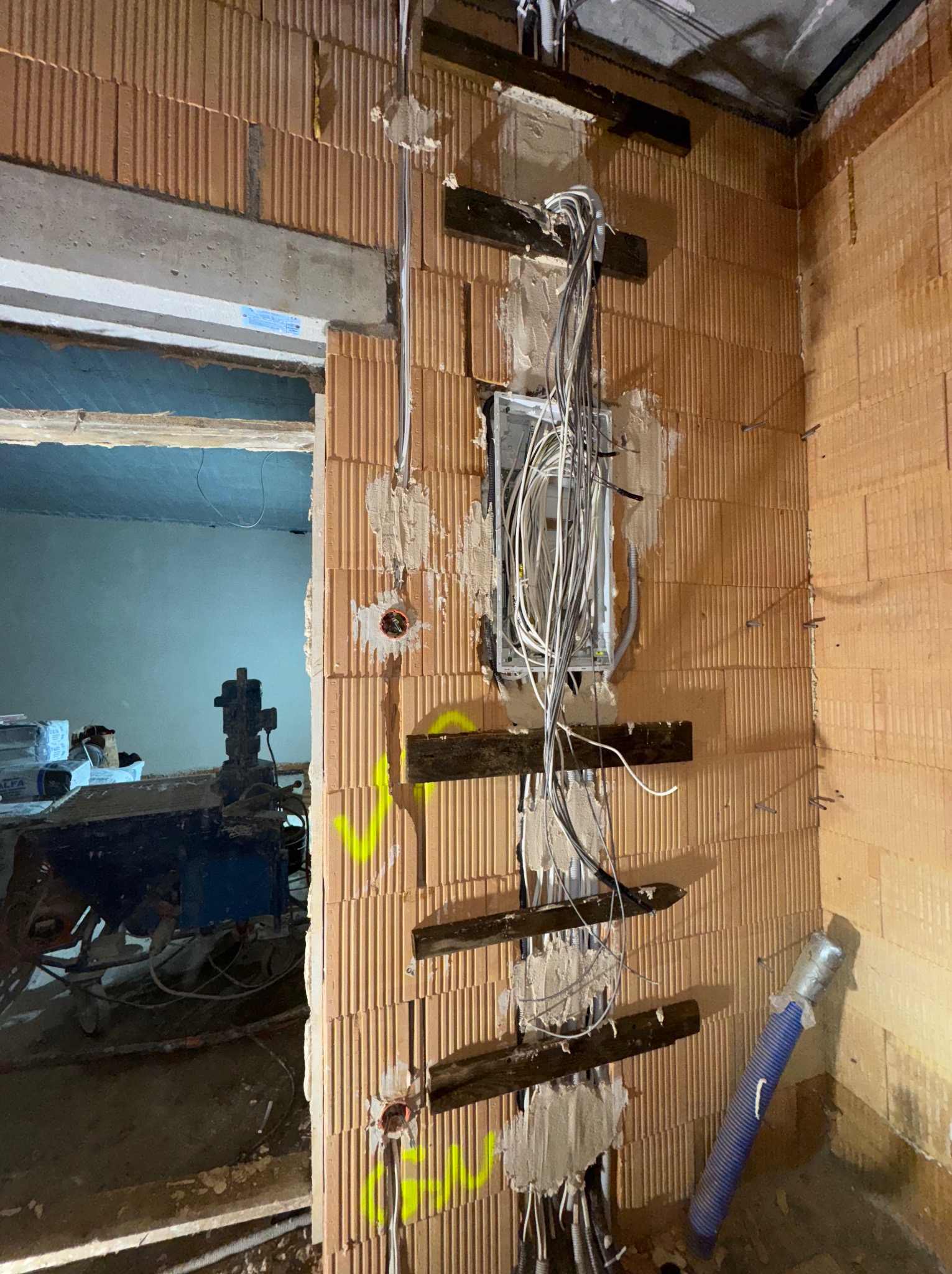 Wewnętrzna ściana z pustaków z zamontowaną puszką elektryczną i poprowadzonymi przewodami. Widoczne drewniane elementy mocujące i fragment pomieszczenia obok.