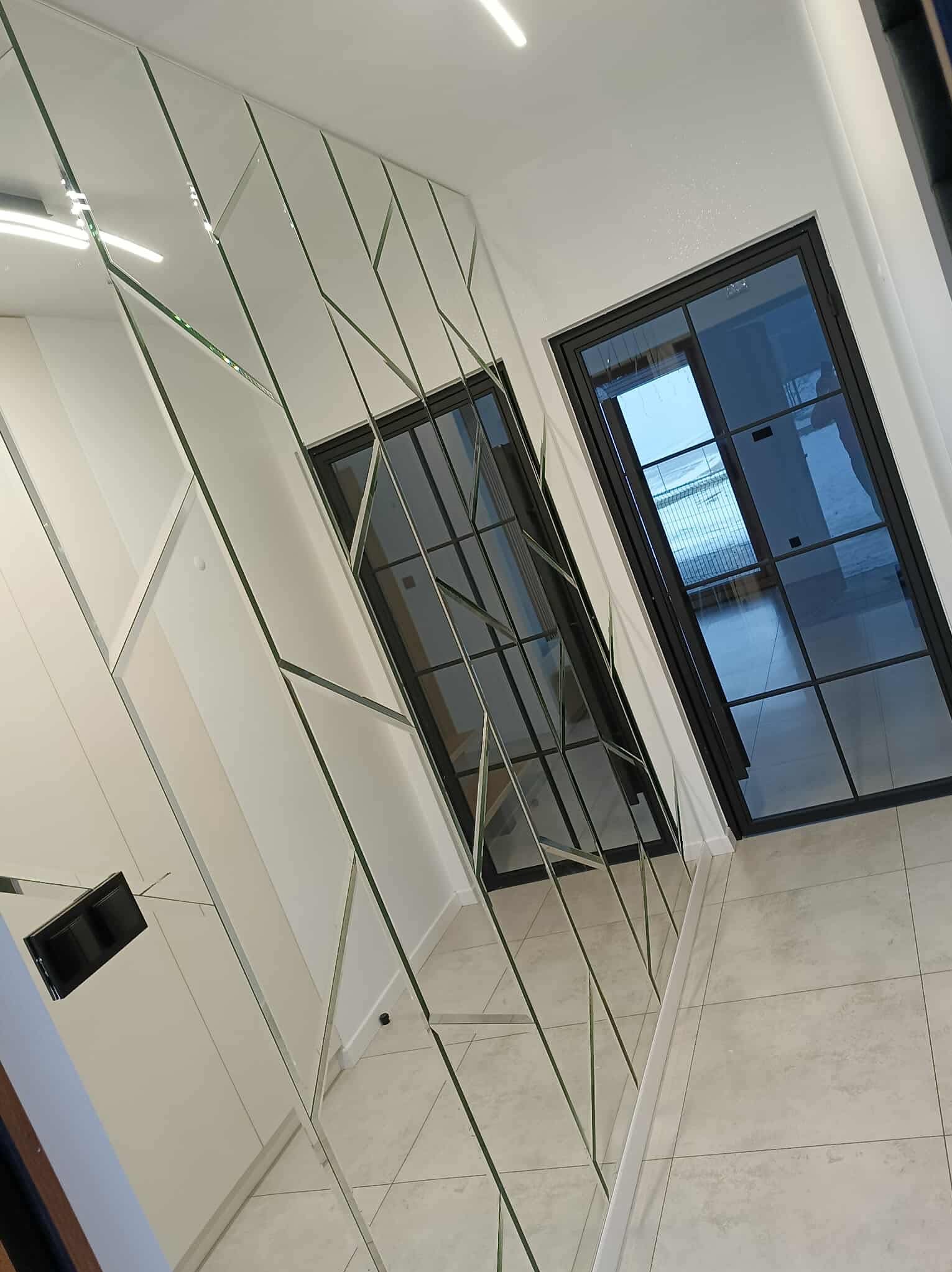 Geometryczna kompozycja z luster na ścianie w korytarzu, odbijająca drzwi z czarnymi szprosami. Nowoczesny design z jasnymi płytkami podłogowymi.