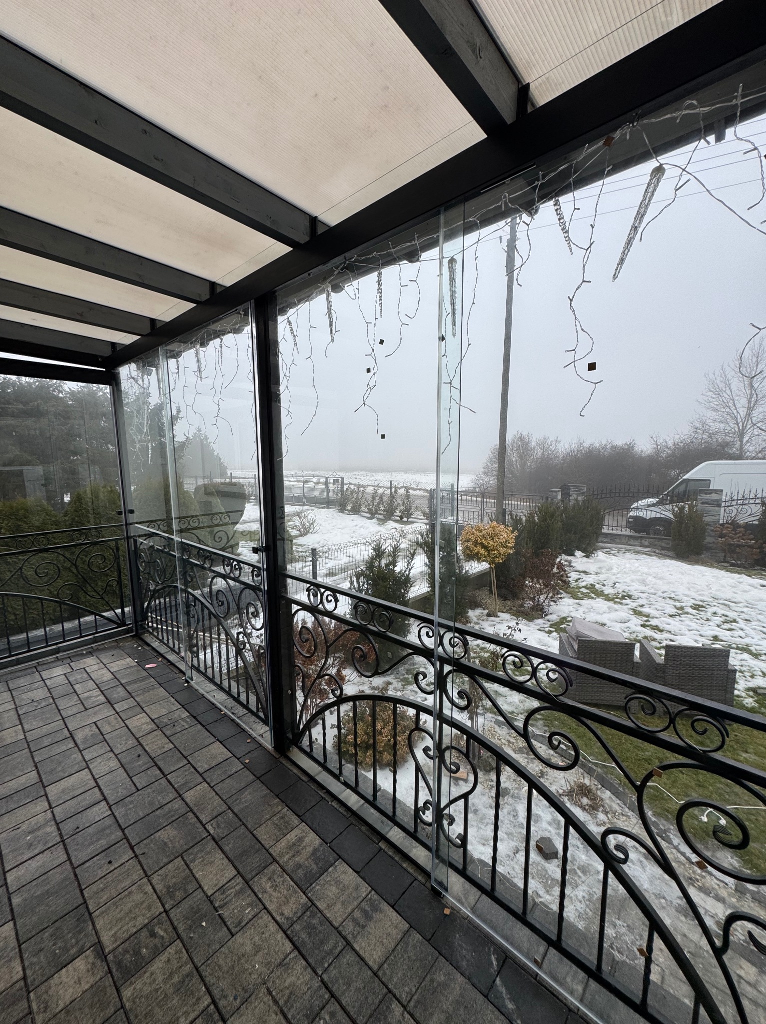 Zadaszony taras z dekoracyjną, kutą balustradą i szklanymi panelami, ozdobiony lampkami. W tle zimowy krajobraz z zaśnieżonym ogrodem i mgłą.
