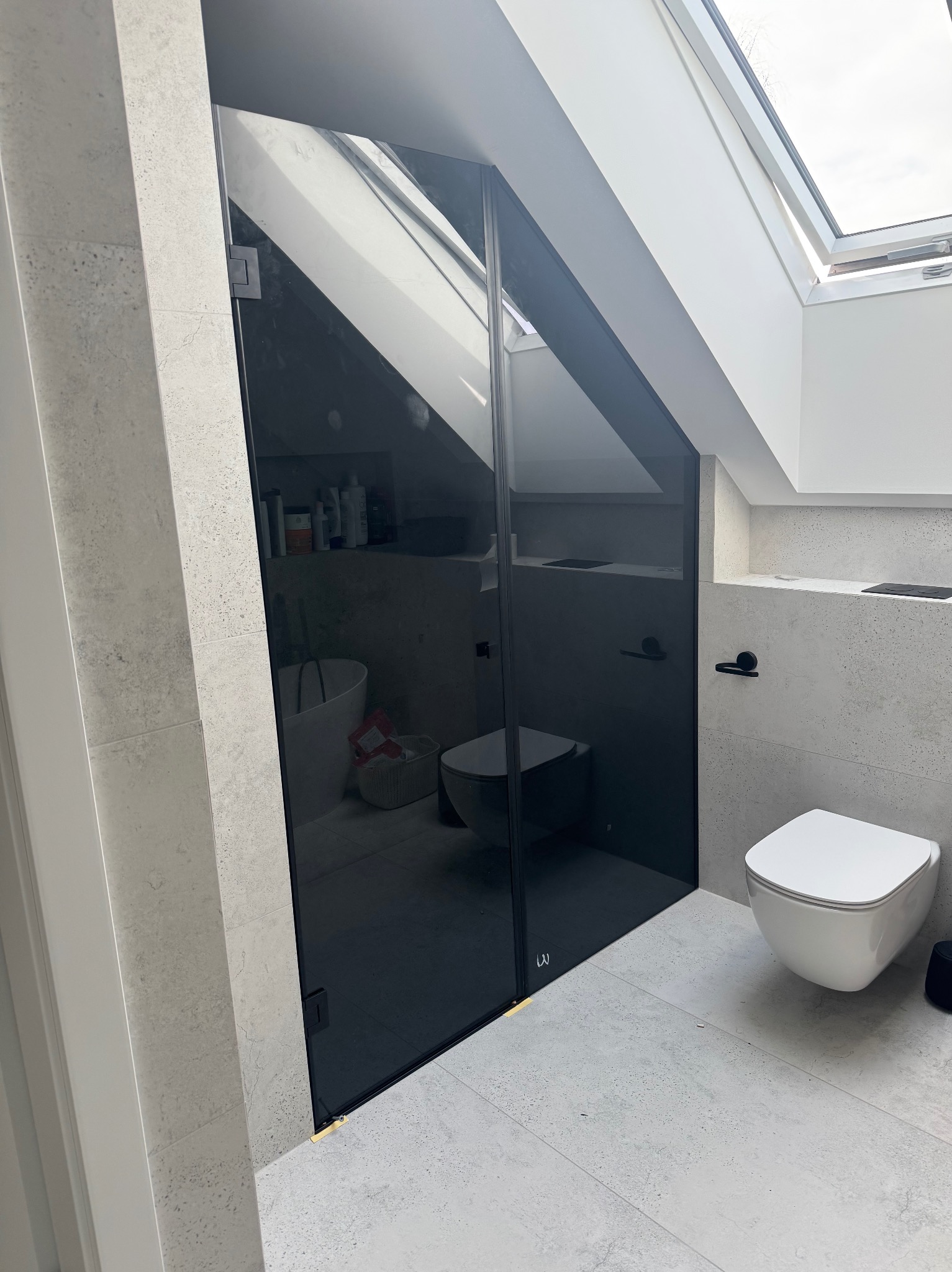 Nowoczesna kabina prysznicowa z czarnego szkła w łazience na poddaszu, widok na toaletę i okno dachowe. Minimalistyczny design, szare płytki.