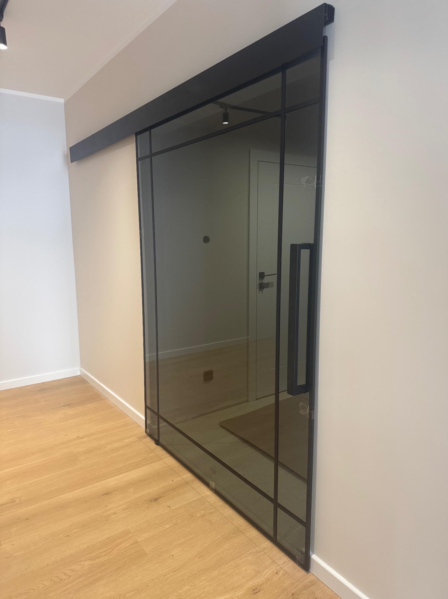 Przesuwne drzwi szklane w czarnej ramie, z widocznym uchwytem, zamontowane w nowoczesnym wnętrzu z drewnianą podłogą. Widok na sąsiednie pomieszczenie.