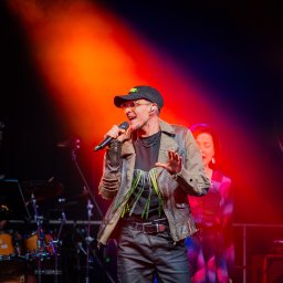 Szkatulski_Photography - Wokalista w skórzanej kurtce, czapce i okularach śpiewa do mikrofonu na scenie oświetlonej czerwonym światłem. W tle widoczna perkusja i postać kobiety.