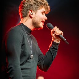 Szkatulski_Photography - Piosenkarz z mikrofonem na scenie w czerwonym oświetleniu. Widoczny srebrny naszyjnik z symbolem wilka. W tle rozmyta postać kobiety.