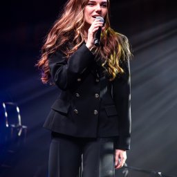 Szkatulski_Photography - Piosenkarka w czarnym garniturze, z mikrofonem na scenie podczas występu. Długie, falowane włosy, profesjonalny makijaż. Scena oświetlona reflektorami.