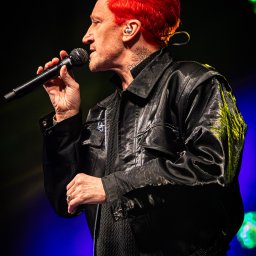 Szkatulski_Photography - Profil wokalisty z czerwonymi włosami, śpiewającego do mikrofonu na scenie. Widoczne tatuaże na szyi, błyszcząca kurtka i dżinsy. Scena oświetlona niebieskim i zielonym światłem.