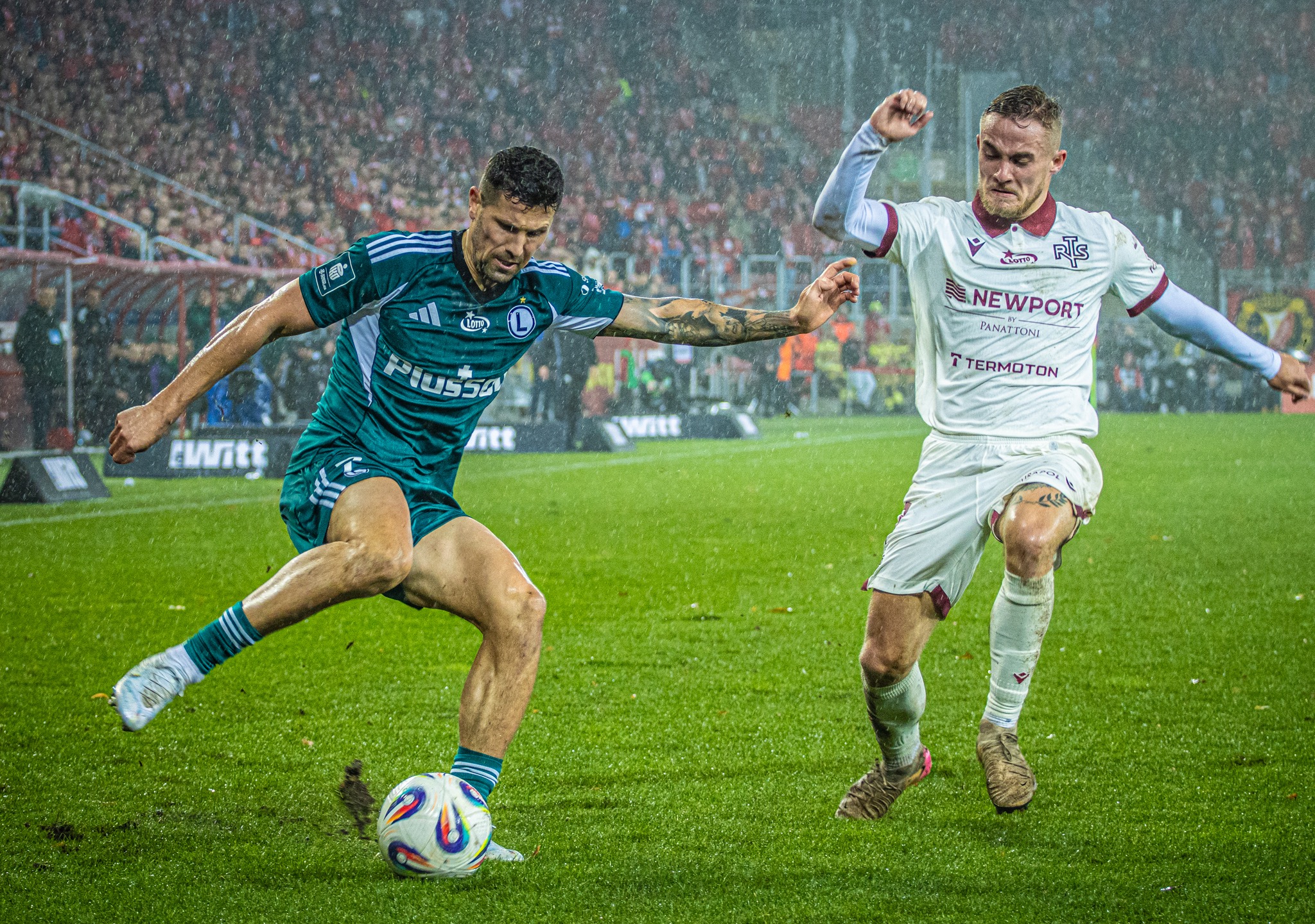 Dynamiczne ujęcie piłkarzy w akcji podczas meczu w deszczu. Jeden z graczy w zielonym stroju kopie piłkę, drugi w białym próbuje go powstrzymać. Tłum kibiców w tle.