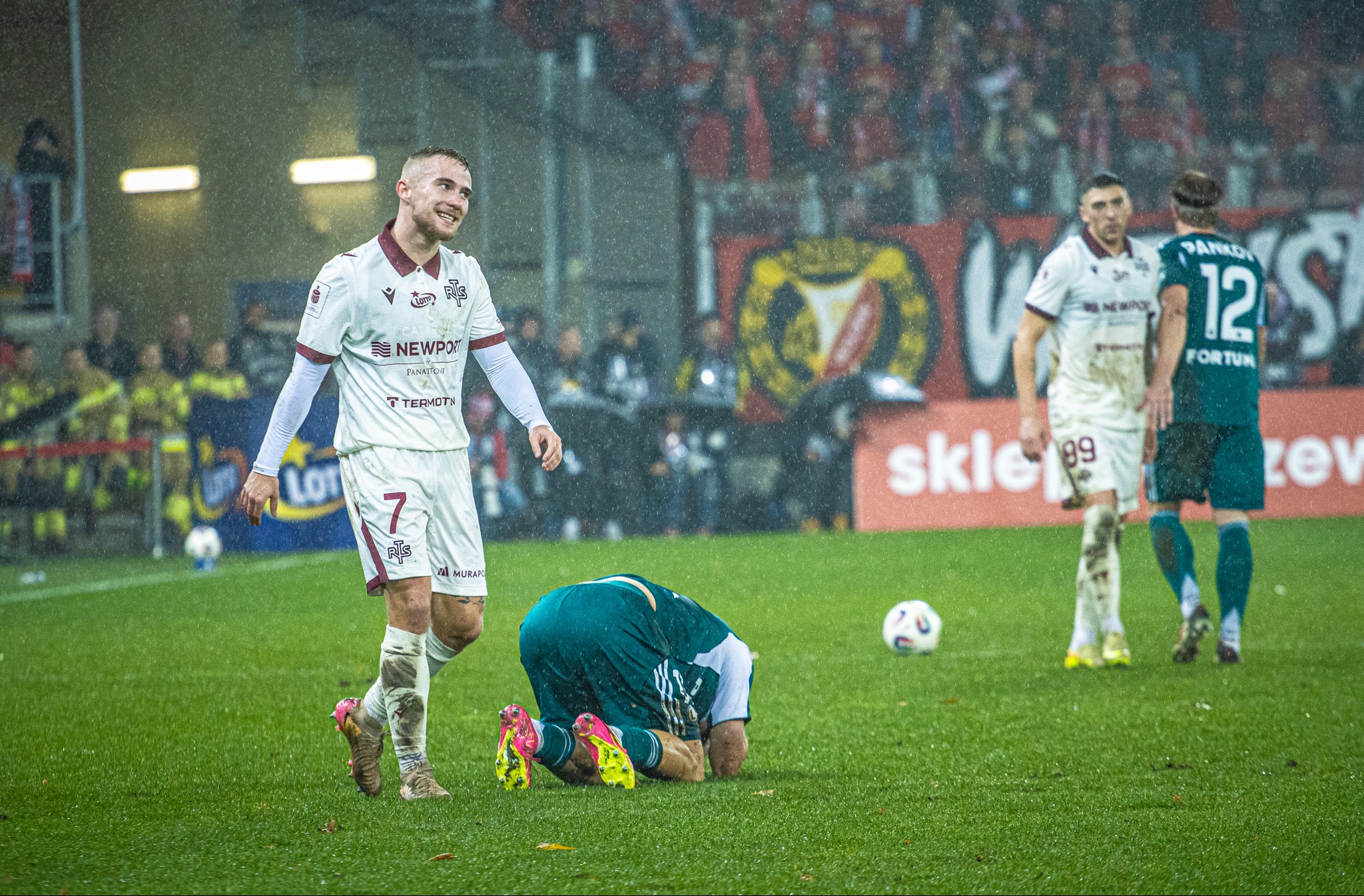 Dynamiczne ujęcie z meczu piłki nożnej w Sulejowie podczas deszczu. Widoczni zawodnicy w akcji, jeden z nich z uśmiechem, inny klęczy na murawie. Intensywne emocje sportowe.