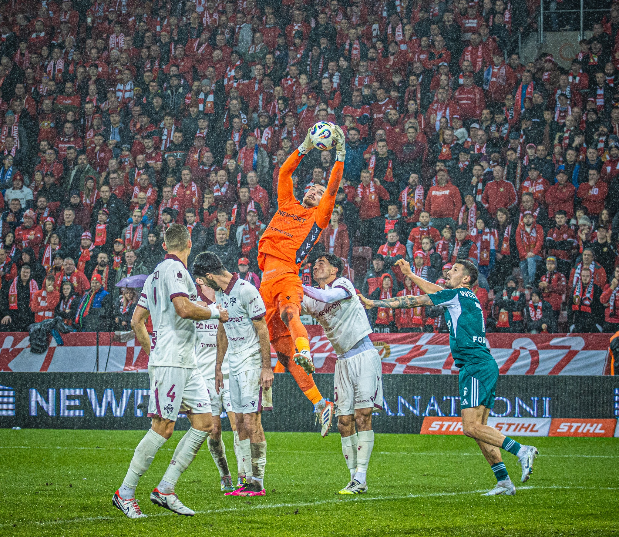 Mecz piłki nożnej w deszczu: bramkarz w pomarańczowym stroju łapie piłkę w powietrzu, otoczony przez zawodników i kibiców na trybunach. Dynamiczne ujęcie.