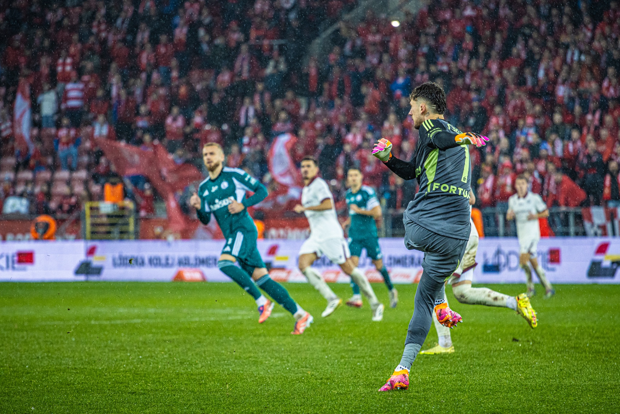 Dynamiczne ujęcie bramkarza w akcji podczas meczu piłki nożnej, z widocznymi zawodnikami w tle i kibicami na trybunach. Deszczowa pogoda dodaje dramatyzmu.