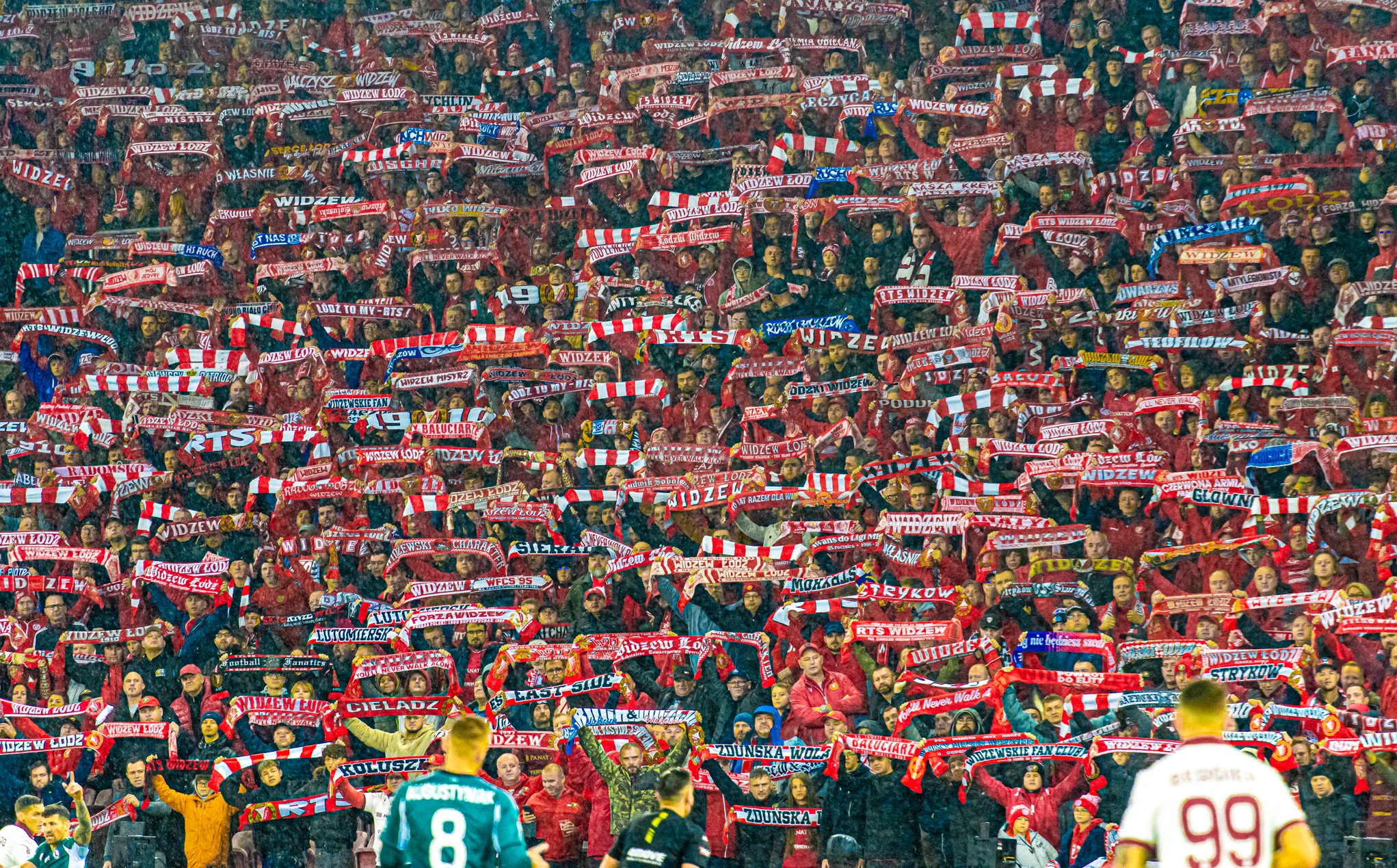 Tłum kibiców Widzewa Łódź na trybunach stadionu, trzymających szaliki w barwach klubowych, tworzących morze czerwieni i bieli. Widoczni piłkarze w dolnej części kadru.