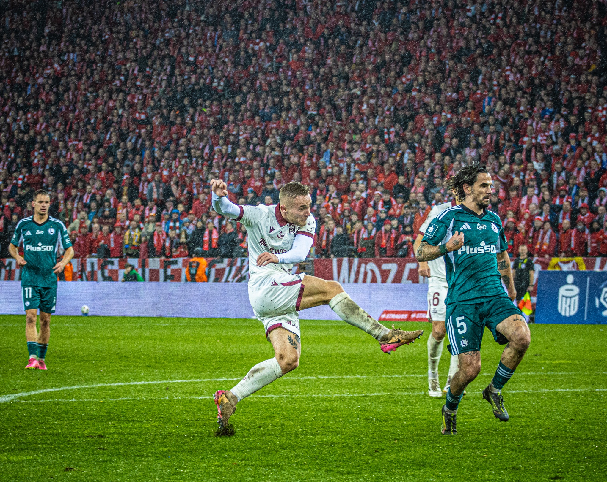 Dynamiczne ujęcie piłkarza w akcji podczas meczu na tle trybun wypełnionych kibicami w deszczowy dzień. Widoczne detale strojów i piłki.