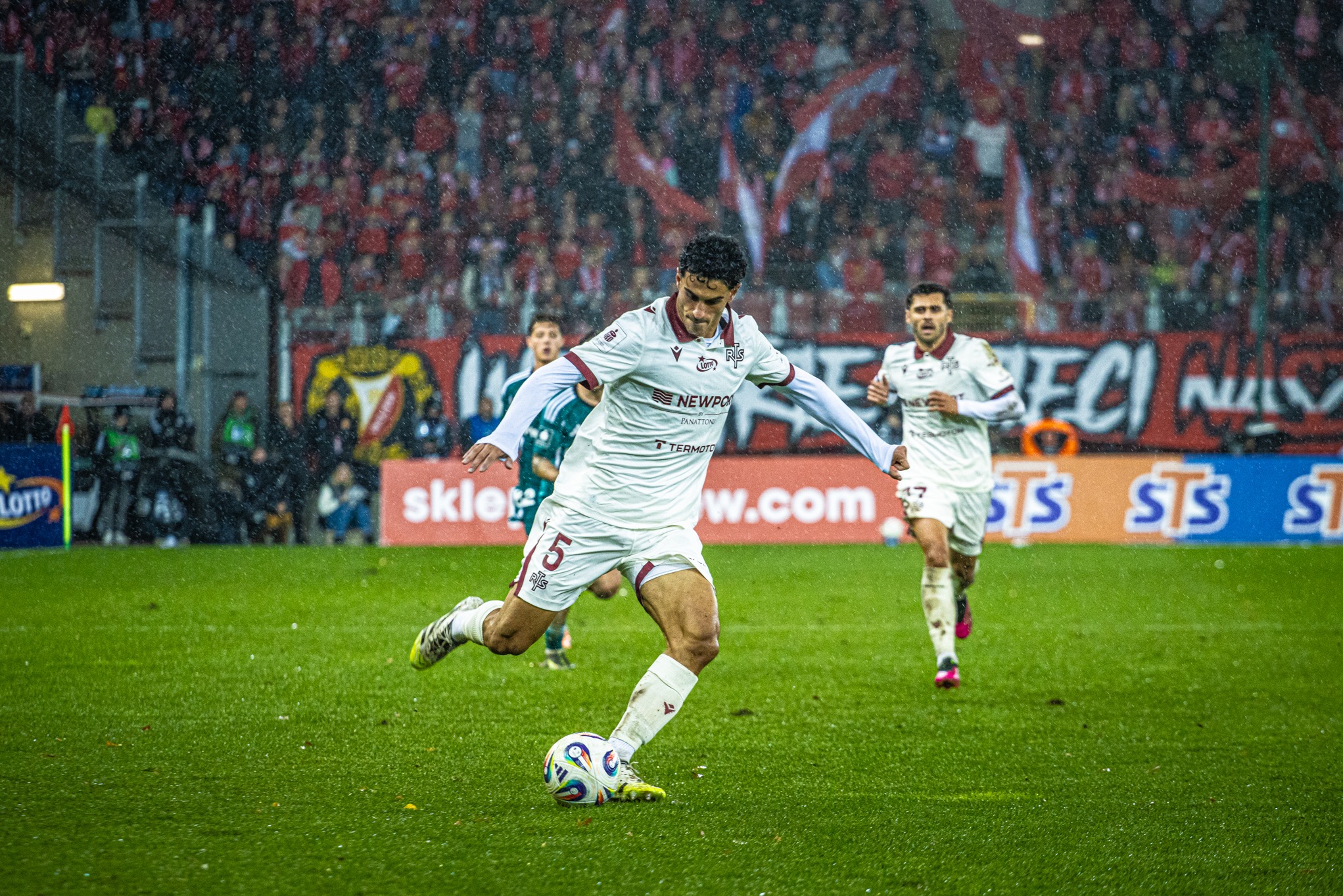 Dynamiczne ujęcie piłkarza w białym stroju podczas meczu na zielonej murawie, na tle trybun wypełnionych kibicami w deszczowy dzień. Widoczne detale stroju i piłki.