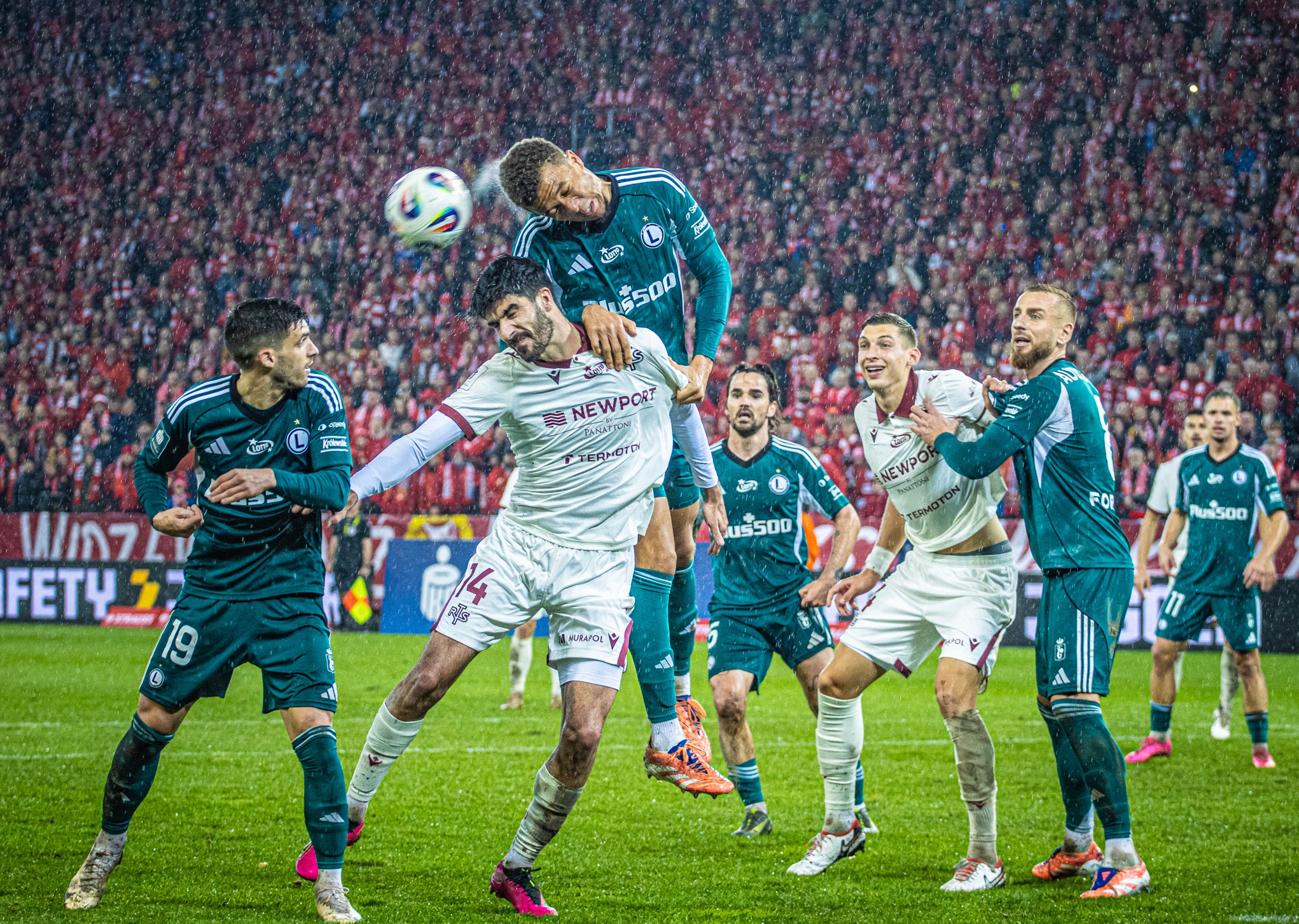 Dynamiczne ujęcie meczu piłki nożnej w deszczu: zawodnicy w akcji, piłka w powietrzu, tłum kibiców na trybunach w tle. Intensywne emocje i sportowa rywalizacja.