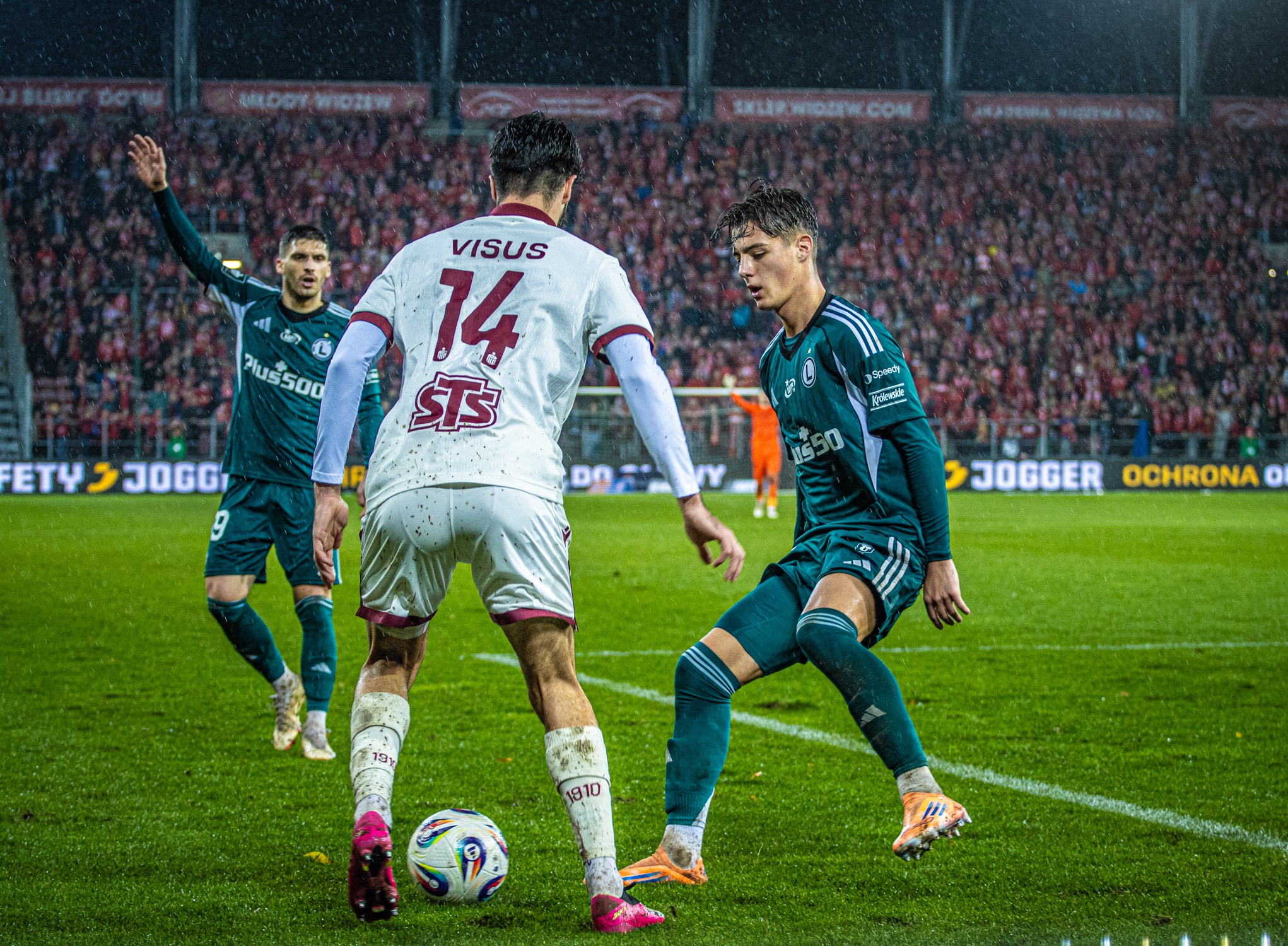 Mecz piłki nożnej: zawodnik w białej koszulce z numerem 14 walczy o piłkę z zawodnikiem w zielonym stroju na tle trybun pełnych kibiców podczas deszczu.