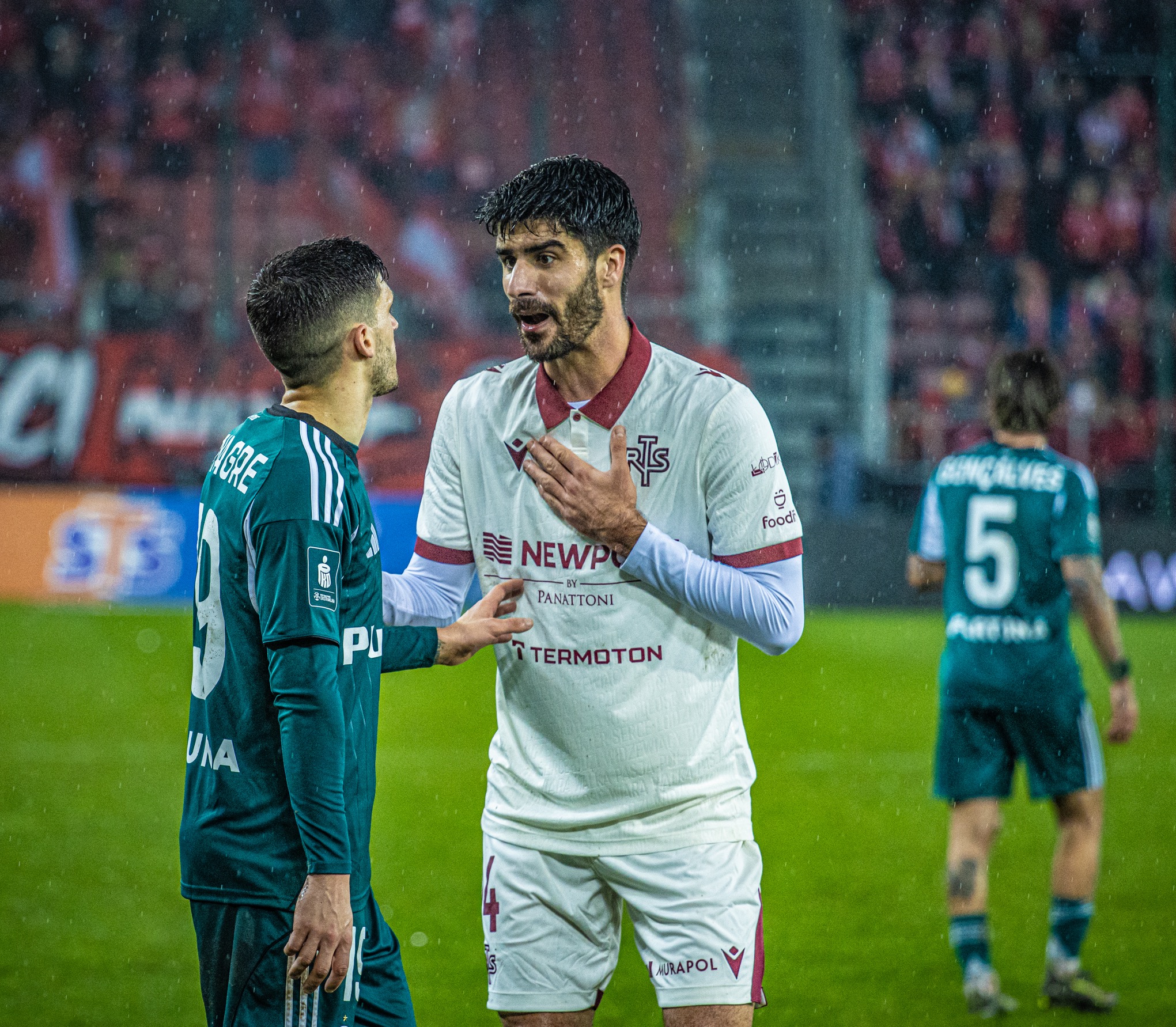 Dwóch piłkarzy w trakcie meczu, jeden w białej koszulce gestykuluje, drugi w zielonej słucha. W tle rozmazane trybuny i padający deszcz.