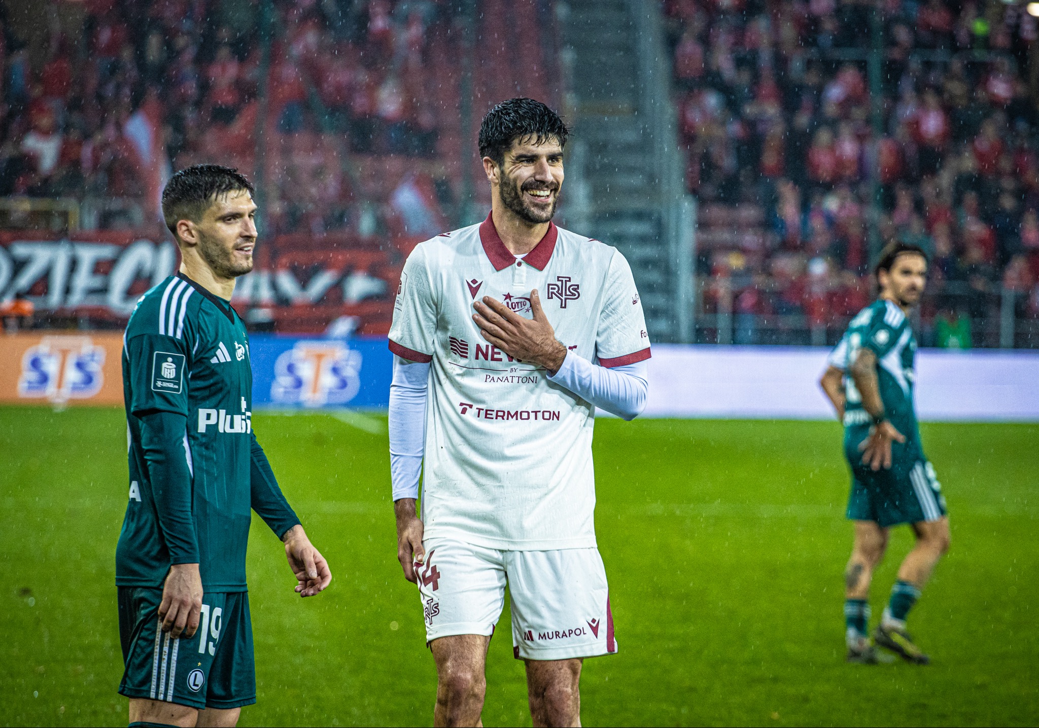 Uśmiechnięty piłkarz w białej koszulce z herbem drużyny na mokrym boisku, w tle kibice i padający deszcz. Ujęcie z meczu.