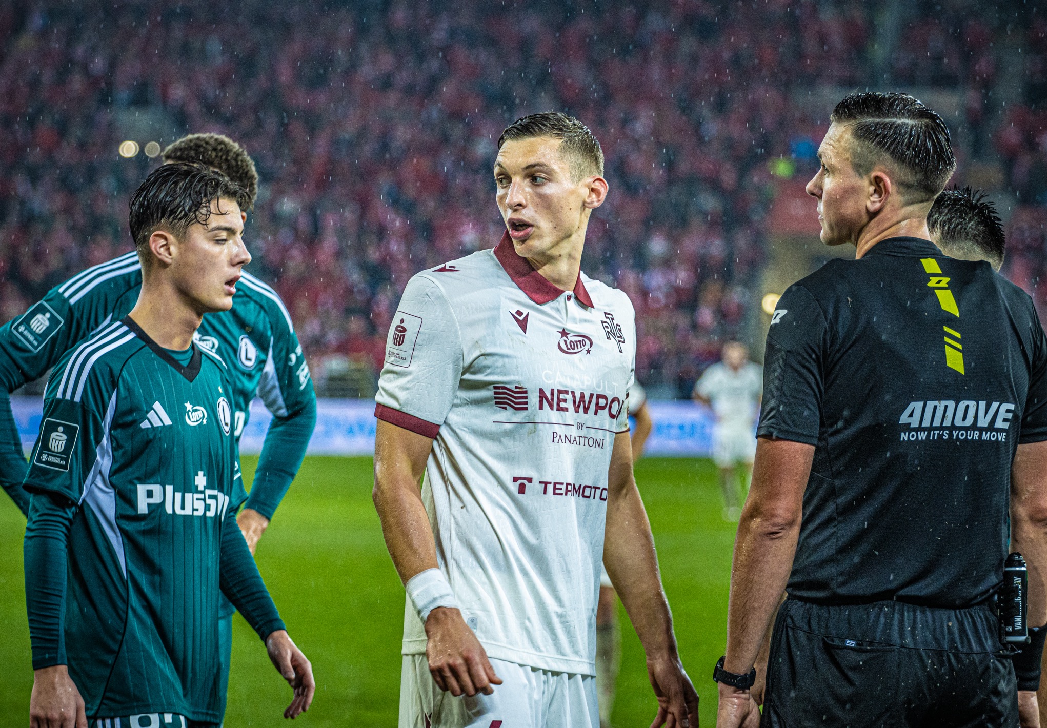 Mecz piłki nożnej w deszczu: zawodnicy w zielonych i białych strojach rozmawiają z sędzią w czarnym uniformie. Widoczny tłum kibiców na trybunach.