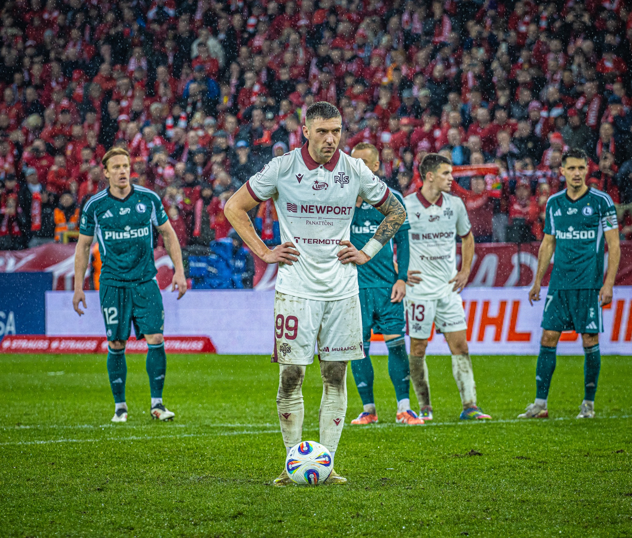 Ujęcie piłkarza z numerem 99 na mokrym boisku, w tle kibice. Zawodnik w białym stroju z logotypami sponsorów, stoi z piłką u stóp. Deszcz pada, tworząc dynamiczny efekt.