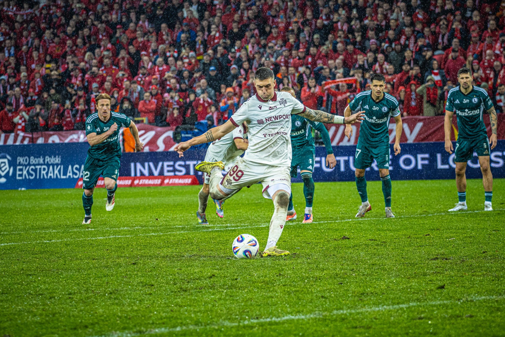 Mecz piłki nożnej w deszczu: zawodnik w białym stroju kopie piłkę, w tle kibice w czerwieni i zawodnicy w zielonych strojach. Dynamiczne ujęcie z bliska.