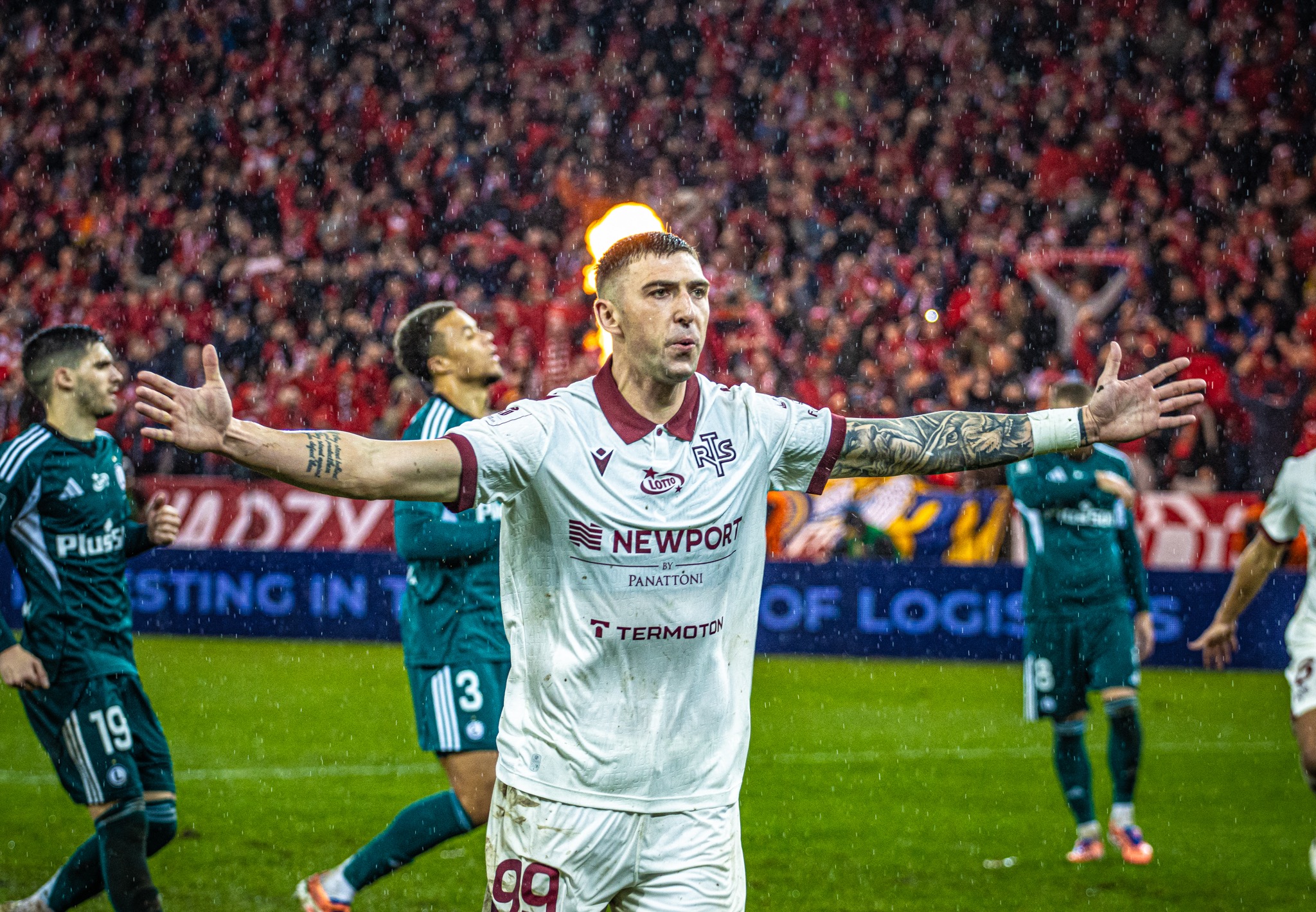 Mokry piłkarz w białej koszulce z rozłożonymi rękami na tle kibiców podczas meczu. Widoczne tatuaże na rękach i krople deszczu. Intensywne oświetlenie.