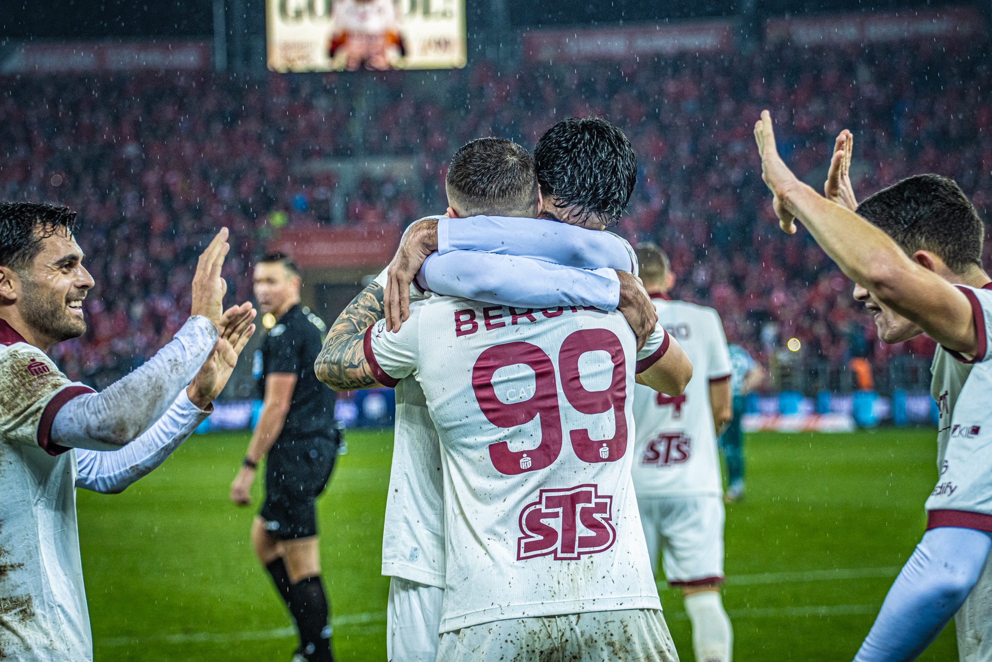 Radosna celebracja piłkarzy w deszczu, z numerem 99 na koszulce, na tle czerwonej publiczności i zielonej murawy. Dynamiczne ujęcie z meczu.