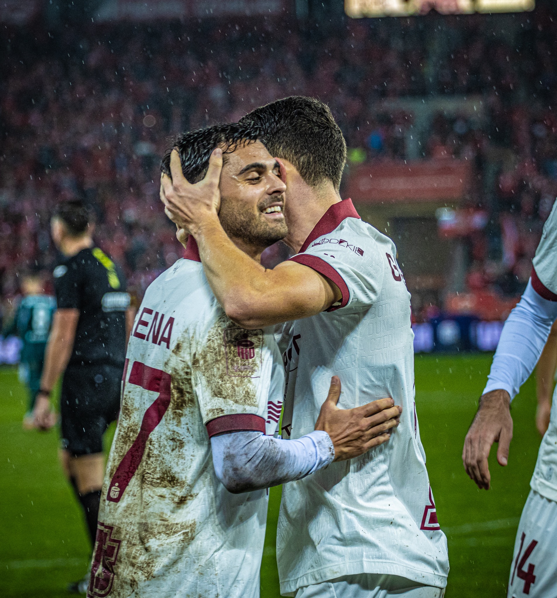 Uśmiechnięci, objęci piłkarze w błotnistych strojach po meczu, na tle czerwonych trybun i padającego deszczu. Emocje sportowe, radość zwycięstwa.