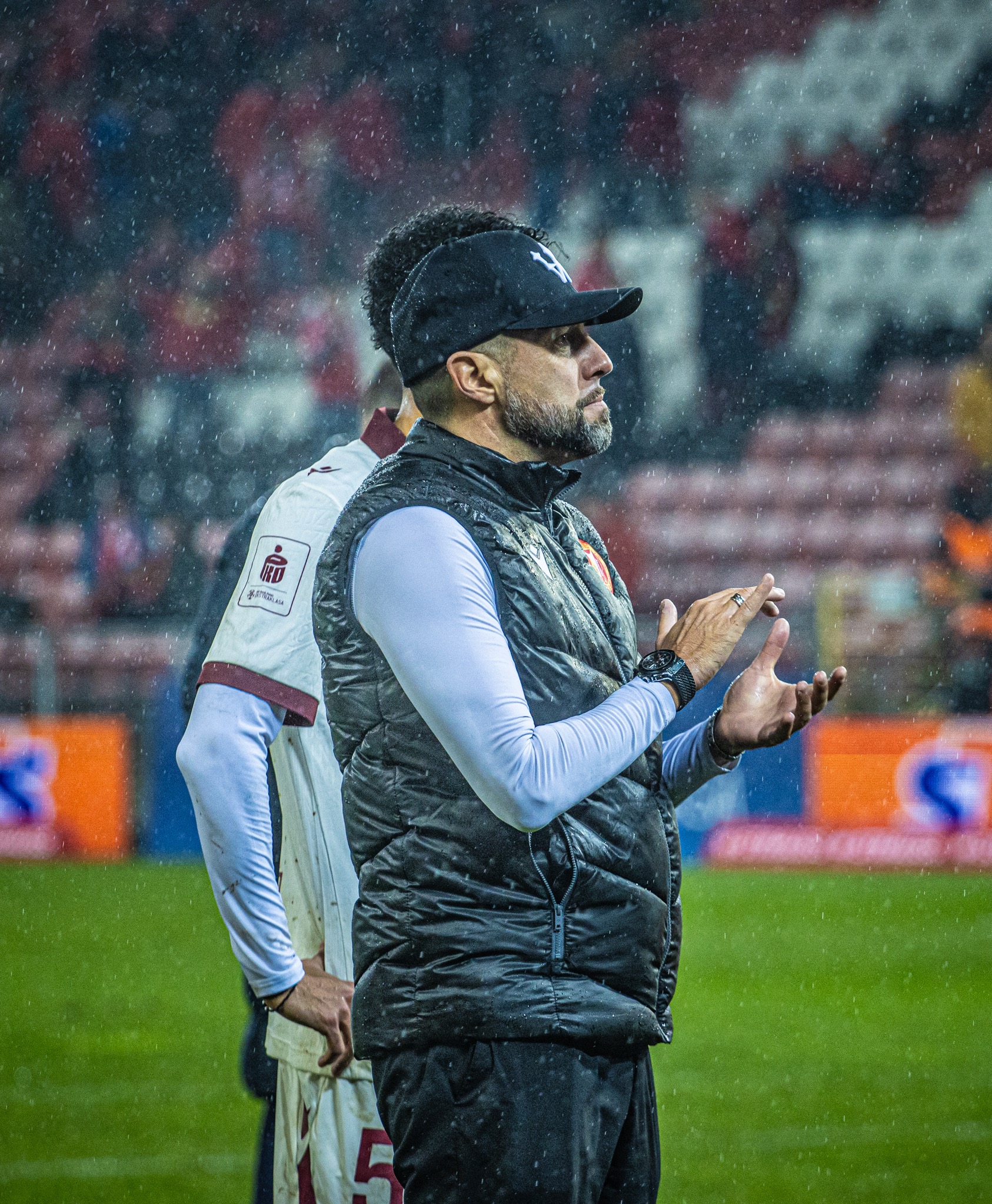 Trener w czapce i kamizelce, klaszcze w deszczu na tle stadionu. Ujęcie z dołu, podkreślające determinację i emocje podczas wydarzenia sportowego.