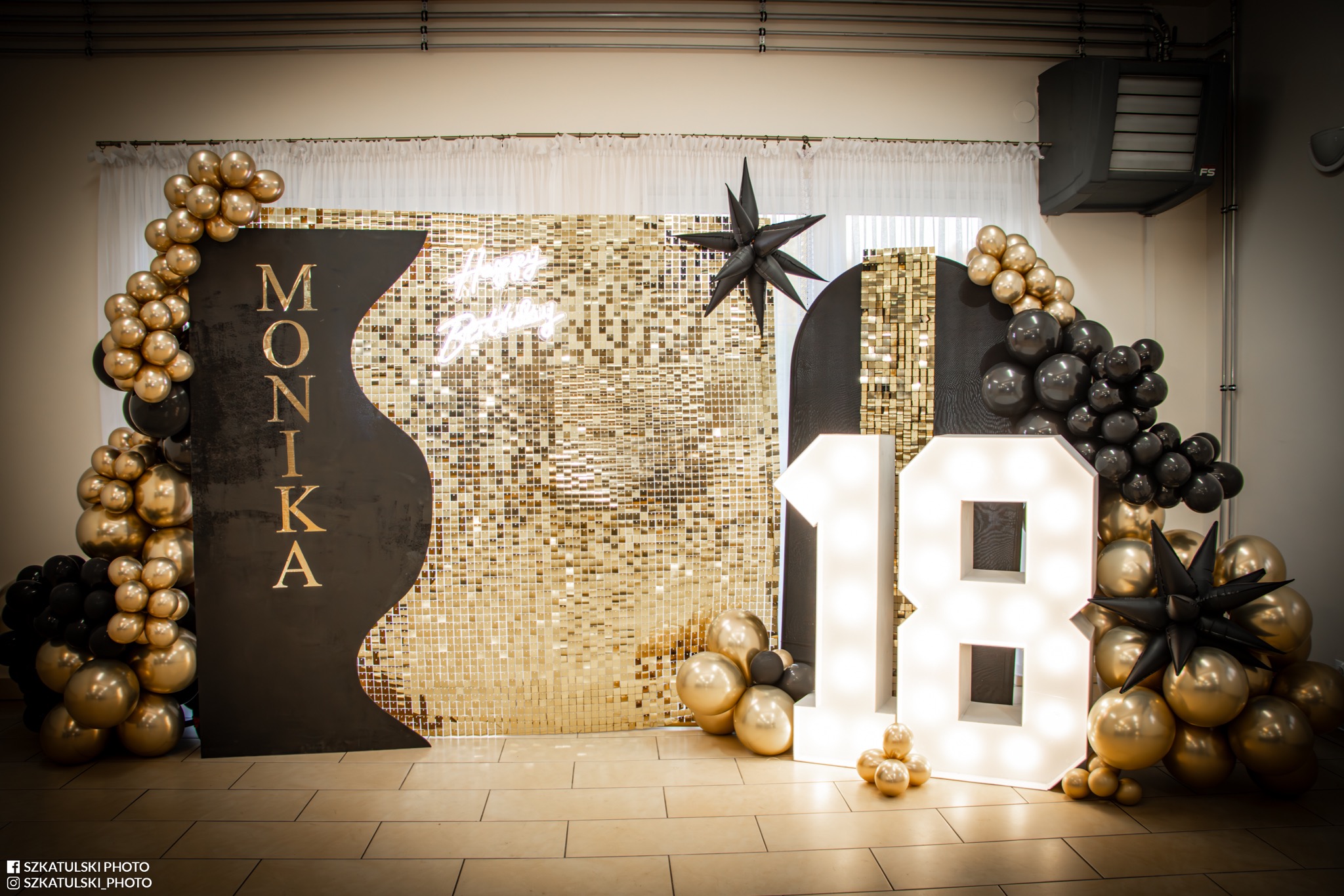 Dekoracja urodzinowa z błyszczącym tłem, balonami w kolorach złota i czerni, oraz podświetlanym napisem '18' i imieniem 'Monika'.