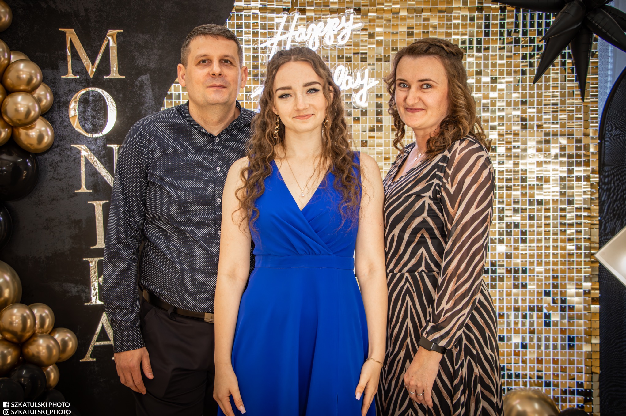 Portret rodzinny: rodzice z córką w niebieskiej sukience pozują na tle dekoracji z balonów i błyszczącej ściany z napisem 'Happy Birthday'.